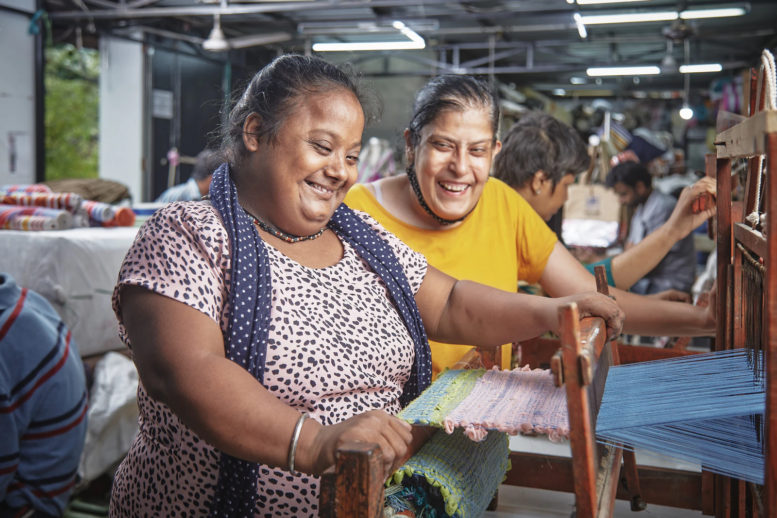 Two smiling women working together at a wooden hand loom, weaving a colourful rug in a busy workshop